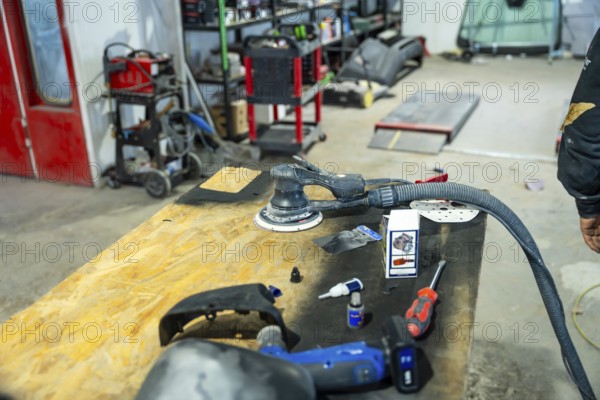Orbital sander, hand tools, and car parts sitting on a wooden workbench inside a busy automotive paint and body repair shop, ready for refinishing work