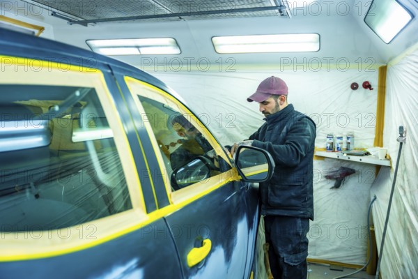 Auto repair technician carefully applying masking tape to a car door and window, protecting parts before professional painting in a clean spray booth workshop, ensuring a precise finish