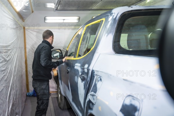 Mechanic applying masking tape to a car surface, meticulously preparing the vehicle for a fresh paint application inside a professional automotive spray booth
