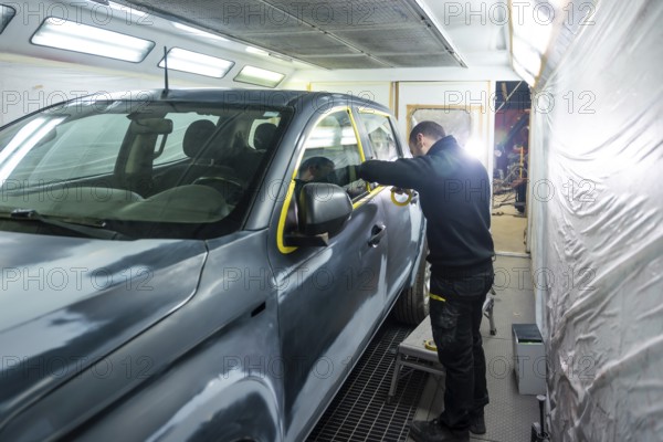 Automotive technician preparing a vehicle for painting by carefully applying protective masking tape around windows and trim in a bright auto body repair booth