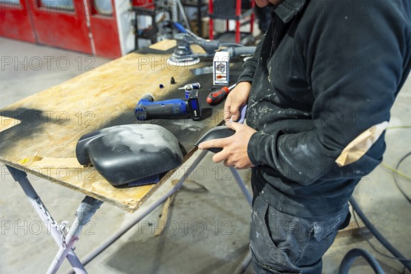 Auto body repairman meticulously sanding a car part, preparing surfaces for paint spraying, restoring vehicle bodywork in a professional automotive workshop with tools on a wooden table