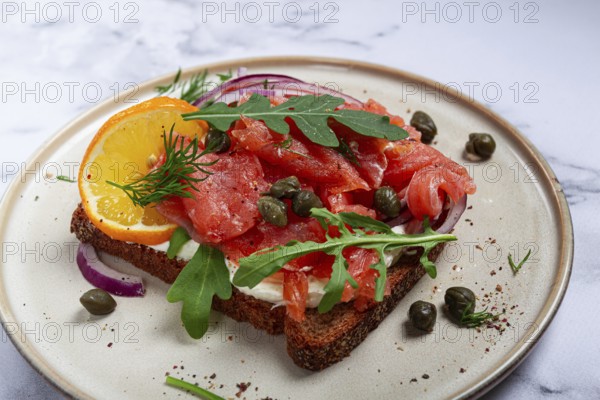 Sandwich with freshly smoked salmon, on black bread, with cottage cheese, with herbs and red onions, capers and pieces of citrus fruits, on a marble countertop, breakfast, no people