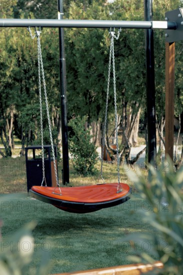 A bright orange hanging swing is set in a serene garden surrounded by trees and grass. The peaceful environment invites relaxation and enjoyment on a sunny afternoon