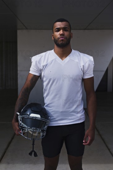 A focused American football player stands outdoors holding his helmet, wearing a white jersey. His determined expression embodies the spirit of the sport