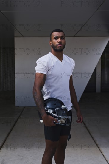 A confident American football player stands in a white jersey, holding a helmet, ready for the game. The athlete exudes determination and strength