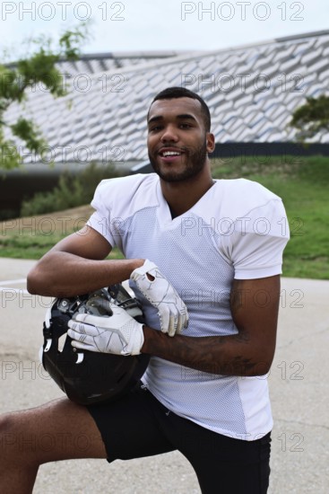 A confident American football player in uniform poses outdoors, holding his helmet. The setting suggests a practice session or casual team meet up with modern architecture