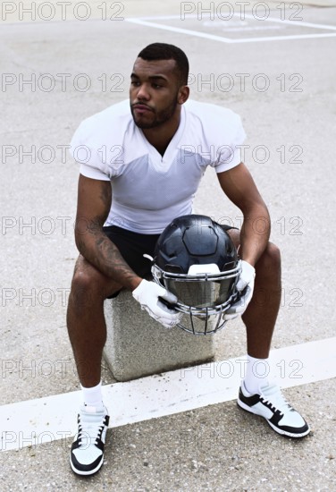 An american football player in a white jersey sits on a bench holding his helmet, reflecting before the game. Focus on determination and readiness in a sports setting