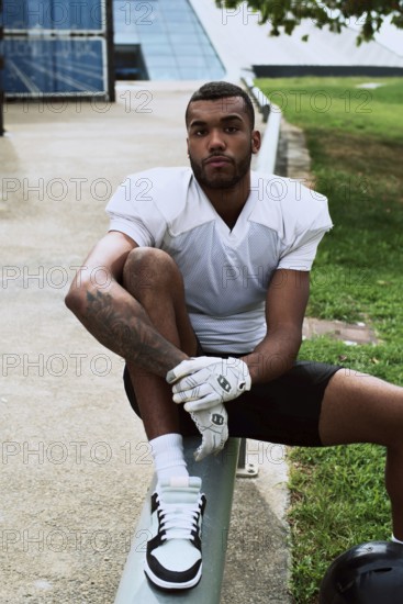 A young American football player in a white jersey sits on a bench outdoors, exuding confidence and readiness. The scene captures the essence of athleticism and determination