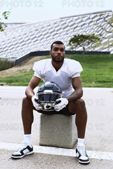 American football player sits on a stone block, holding a helmet. The scene is set in an urban park with modern architecture in the background