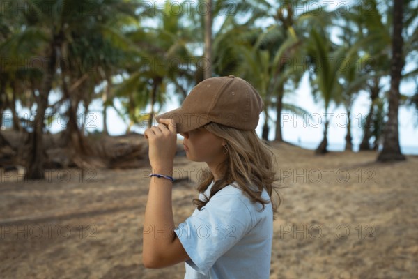 A young woman wearing a casual brown cap and blue shirt stands on a sandy beach, surrounded by lush palm trees, capturing a serene coastal atmosphere in Thailand