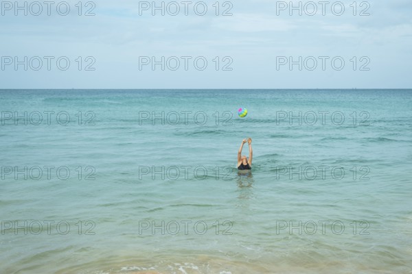 A young woman engaged in playful activity with a colorful ball in the calm ocean water in Thailand, reflecting a summer day joy and relaxation in a vacation environment