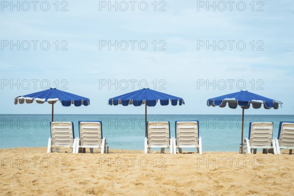 A serene beach scene shows neatly aligned lounge chairs beneath blue umbrellas facing a tranquil sea in Thailand. The golden sand and clear sky complete the idyllic setting