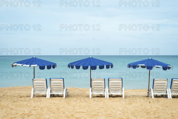 A serene beach scene with three blue umbrellas and white lounge chairs on golden sand facing a calm, expansive ocean in Thailand. The tranquil setting invites relaxation and leisure in vacation