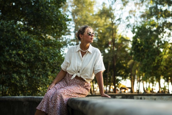 A stylish woman in sunglasses and summer attire sits on a stone ledge surrounded by lush greenery in Thailand. The scene captures a moment of peaceful reflection in vacation