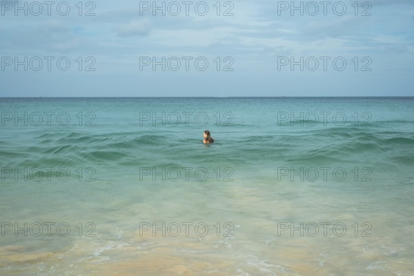 A young woman enjoys a swim in the clear turquoise waters of a tranquil beach in vacation, creating a peaceful scene with gentle waves, sandy shores, and a vast horizon in Thailand