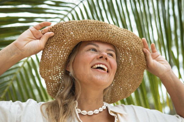 A joyful woman wearing a straw hat and shell necklace smiles under palm leaves, capturing a perfect summer day in Thailand. Her happiness and admiration for nature shine brightly