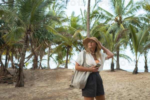 A woman in a summer outfit and hat explores a sandy beach in Thailand, holding her phone under lush palm trees, evoking a sense of adventure and tropical relaxation in vacation