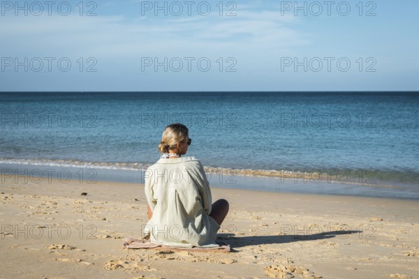 A woman sits calmly on a sandy beach, gazing at the serene ocean under a clear sky in Thailand. Her relaxed posture conveys peace, introspection, and connection with nature in vacation