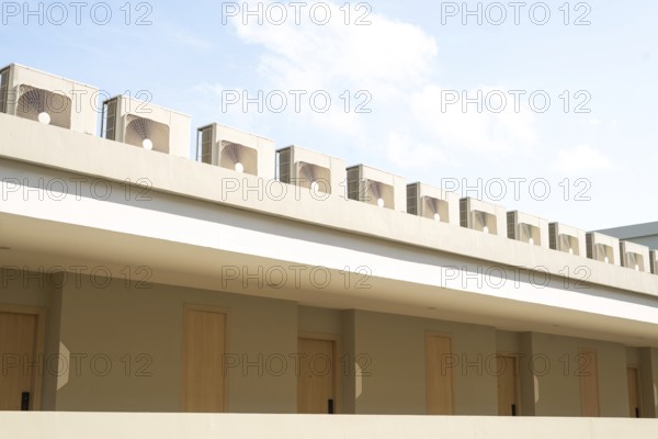 A row of air conditioning units on a modern building rooftop against a clear blue sky in Thailand. The beige exterior with wooden doors creates a minimalist, clean appearance