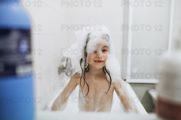 A joyful child is playing with bubbles in a bathtub, with a big smile and wet hair. The scene captures the innocence and fun of bath time in a bright, clean setting