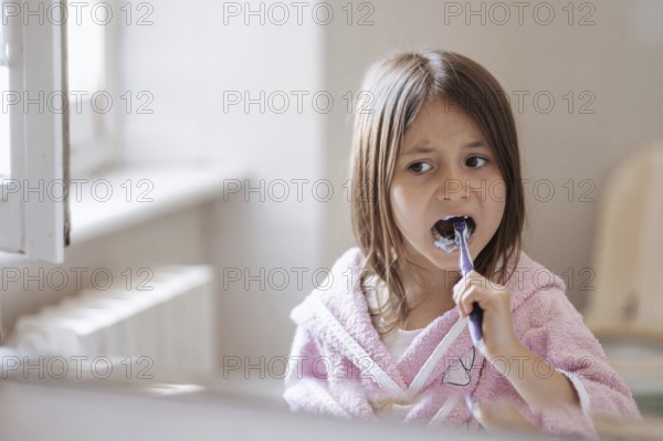 A young girl wearing a pink bathrobe brushes her teeth with a toothbrush, highlighting morning hygiene routine in a bright bathroom setting, promoting dental health