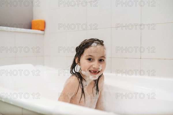 A cheerful child bathes in a white tub, surrounded by soap bubbles and foam. The joy of bath time is evident in the child playful expression and sudsy hair