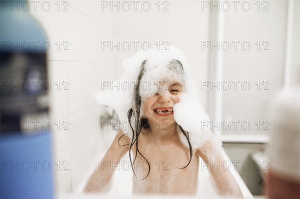 A joyful child enjoys a playful bath, covered in foamy soap bubbles, smiling widely. The bright bathroom setting adds to the cheerful and relaxed atmosphere