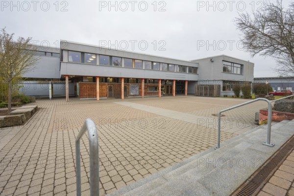 A schoolyard with extensive pavement, gray building structure and outdoor tree, Schlehengäu Grund Schule Gechingen, Calw district, Germany