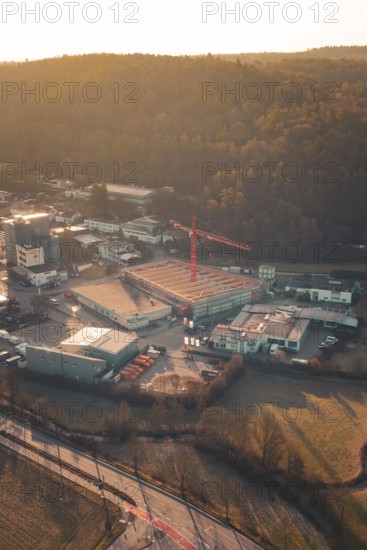 View of an industrial landscape with construction crane and buildings at the foot of wooded hills at sunset, Rewe new building, Aidlingen, Böblingen district, Germany