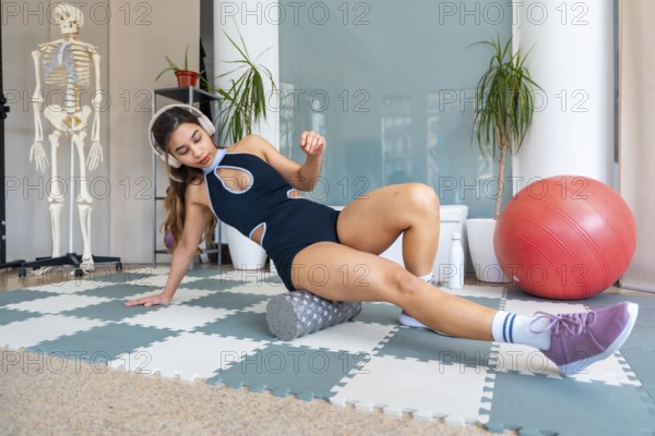 Young woman in activewear using a foam roller on a mat to perform self myofascial release for leg muscles in a clinical recovery and physiotherapy setting, wearing headphones