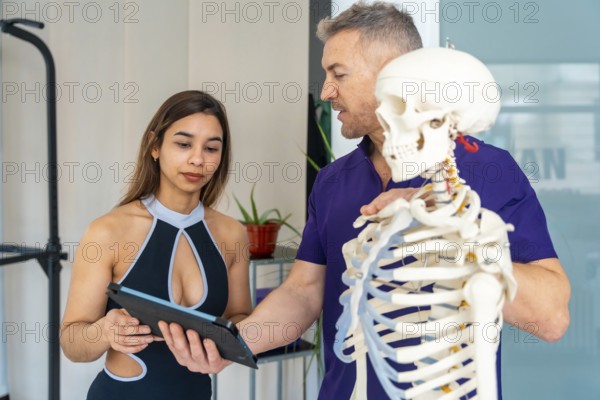 Physiotherapist explains musculoskeletal anatomy using a skeleton model while female patient holds a tablet during a professional clinic consultation for recovery and rehabilitation