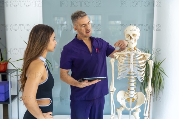 Physiotherapist in purple scrubs pointing at a human skeleton model to a female patient, explaining anatomy during a sports injury recovery session in a modern clinic