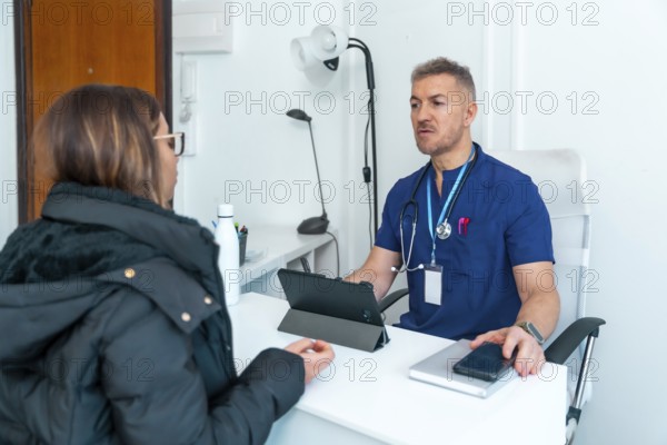 Healthcare professional wearing blue scrubs and a stethoscope talking with a female patient, providing a medical consultation and discussing health concerns in a clean modern clinic room