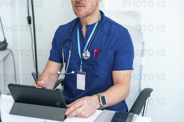 Medical professional in blue scrubs wearing a stethoscope and id card, using a digital tablet while sitting at a white office desk, focusing on digital healthcare tasks