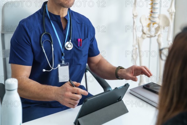 Doctor in scrubs discussing health information with patient, using a tablet for digital records during a clinical appointment, with a skeleton model highlighting anatomical context