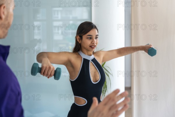 Young woman doing lateral dumbbell raises with a physiotherapist in clinic, focused on shoulder rehabilitation, strength building and guided physical therapy for recovery