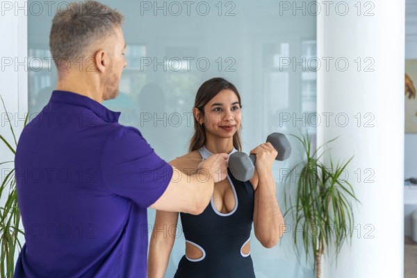 Physiotherapist male trainer assisting a young woman client with rehabilitation and strength training exercises using a dumbbell, focusing on medical recovery and physical therapy in a gym setting