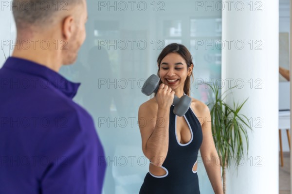 Smiling young woman doing biceps curls with a dumbbell under the guidance of a male physiotherapist, focusing on medical recovery and rehabilitation for strength training and wellness