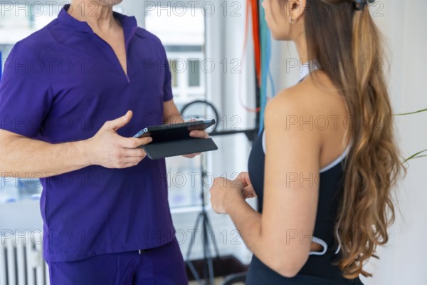 Physiotherapist wearing purple scrubs discussing a medical recovery or sports osteopathy therapy plan with a female patient using a digital tablet during a consultation in a brightly lit clinic