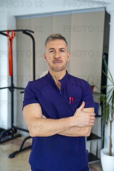 Male physiotherapist in purple uniform stands with arms crossed in a clinic, representing concepts of sports medicine, rehabilitation, and medical recovery