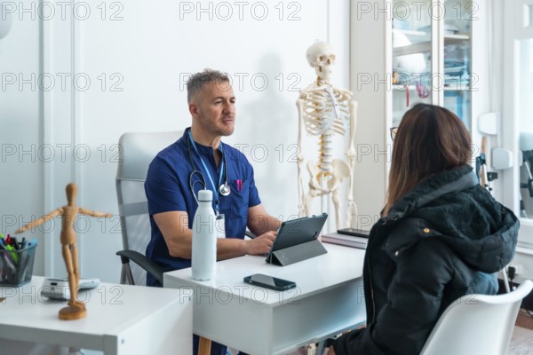 Male doctor in scrubs and stethoscope is talking with a female patient seated across his desk, discussing health concerns during a professional consultation in a medical office with a skeleton model