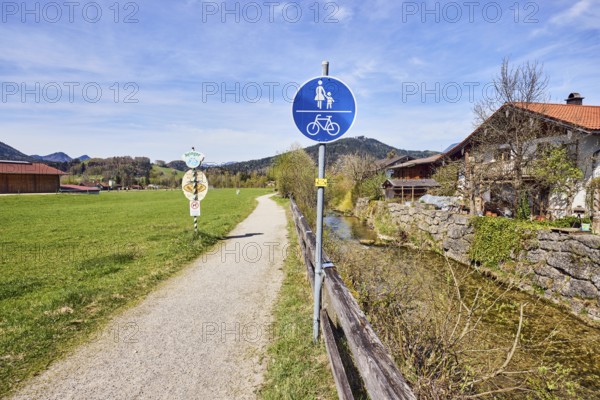 Common footpath and cycle path, traffic sign, footpath and cycle path, river Rote Traun, riverbed, quarry stone wall, wooden fence, meadow, mountains, coniferous forest, trees, bushes, bare winter tree, alpine building, milky blue sky, cumulus clouds, cirrostratus clouds, Inzell, district of Traunstein, Bavaria, Germany