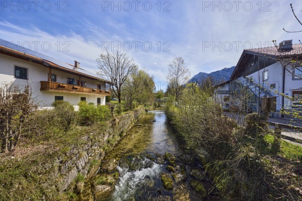 Rote Traun river, fortified bank, riverbed, quarry stone wall, rapids with stones, bushes with spring leaves, shrubs, bare winter trees, houses, alpine buildings, blue sky, cirrus clouds, cirrostratus clouds, Traunsteiner Straße, Inzell, district of Traunstein, Bavaria, Germany