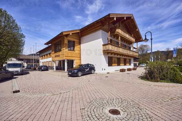 Town hall, commercial buildings, alpine buildings, roofing, balcony, building material wood, lantern, flagpoles, road surface made of concrete paving stones and paving stones, cars, trees, blue sky, cirrus clouds, Rathausplatz square, Inzell, district of Traunstein, Bavaria, Germany