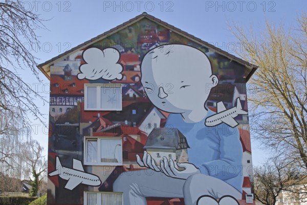 Mural by CASE MacAclaim and Millo, street art, graffiti, child carrying house in hands, illustration, comic, airplane, carrying, Andreas von Chrzanowski, Francesco Camillo Giorgino, house facade, Bad Vilbel, Wetterau, Hesse, Germany