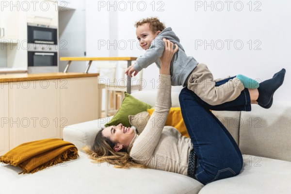 Mother lying on a sofa at home, joyfully playing with her young son, lifting the toddler up in the air with her hands and feet, sharing a happy and loving family moment