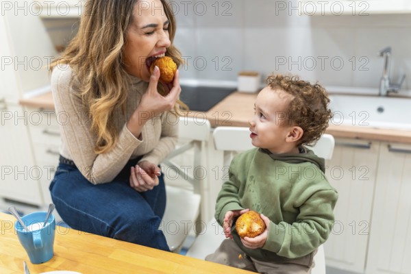 Mother and young son sit at a bright kitchen counter sharing muffins and morning coffee, enjoying a cozy, candid breakfast moment of bonding, joy and domestic togetherness
