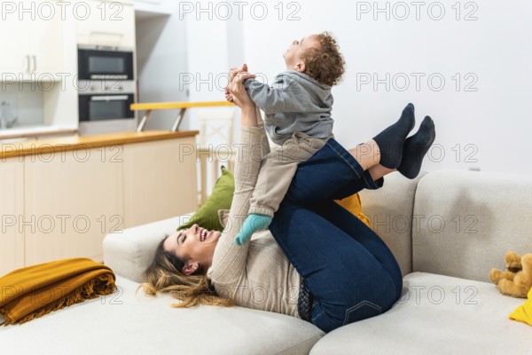 Mother lies on sofa lifting her laughing toddler into the air for a playful airplane ride, sharing a warm, joyful bonding moment of carefree fun and affection at home
