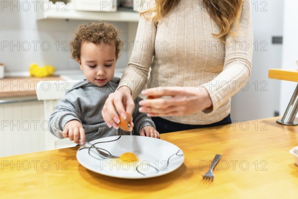 Mother teaching young child to crack an egg and prepare ingredients at the kitchen counter, bonding over baking and meal prep in a warm, casual home setting