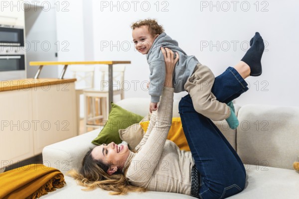 Mother lying on couch lifts smiling toddler in playful airplane pose, sharing a cozy, candid bonding moment of joy and connection in their modern living room